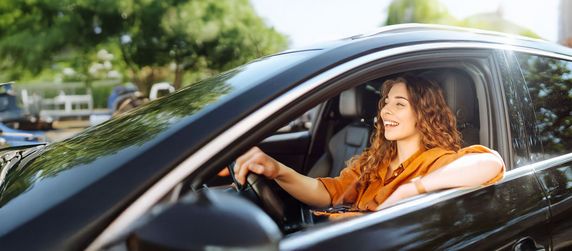 Eine Frau mit lockigem Haar, die ein orangefarbenes Oberteil trägt und ein schwarzes Auto fährt. Sie lächelt und schaut aus dem Fenster.
