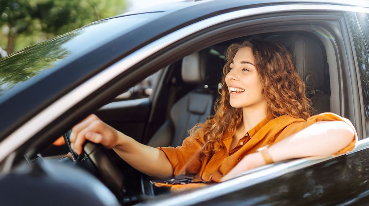 Eine Frau mit lockigem Haar, die ein orangefarbenes Oberteil trägt und ein schwarzes Auto fährt. Sie lächelt und schaut aus dem Fenster.