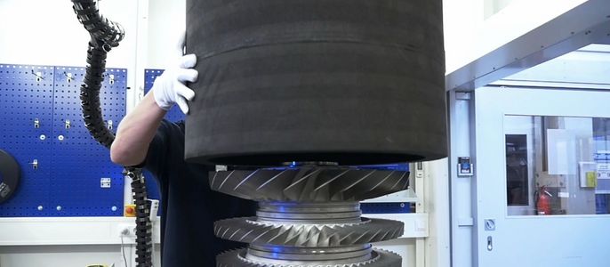 Eine Person in einer Werkstatt, die mit einem großen zylindrischen Gegenstand hantiert, der auf einem metallischen Zahnradmechanismus liegt.