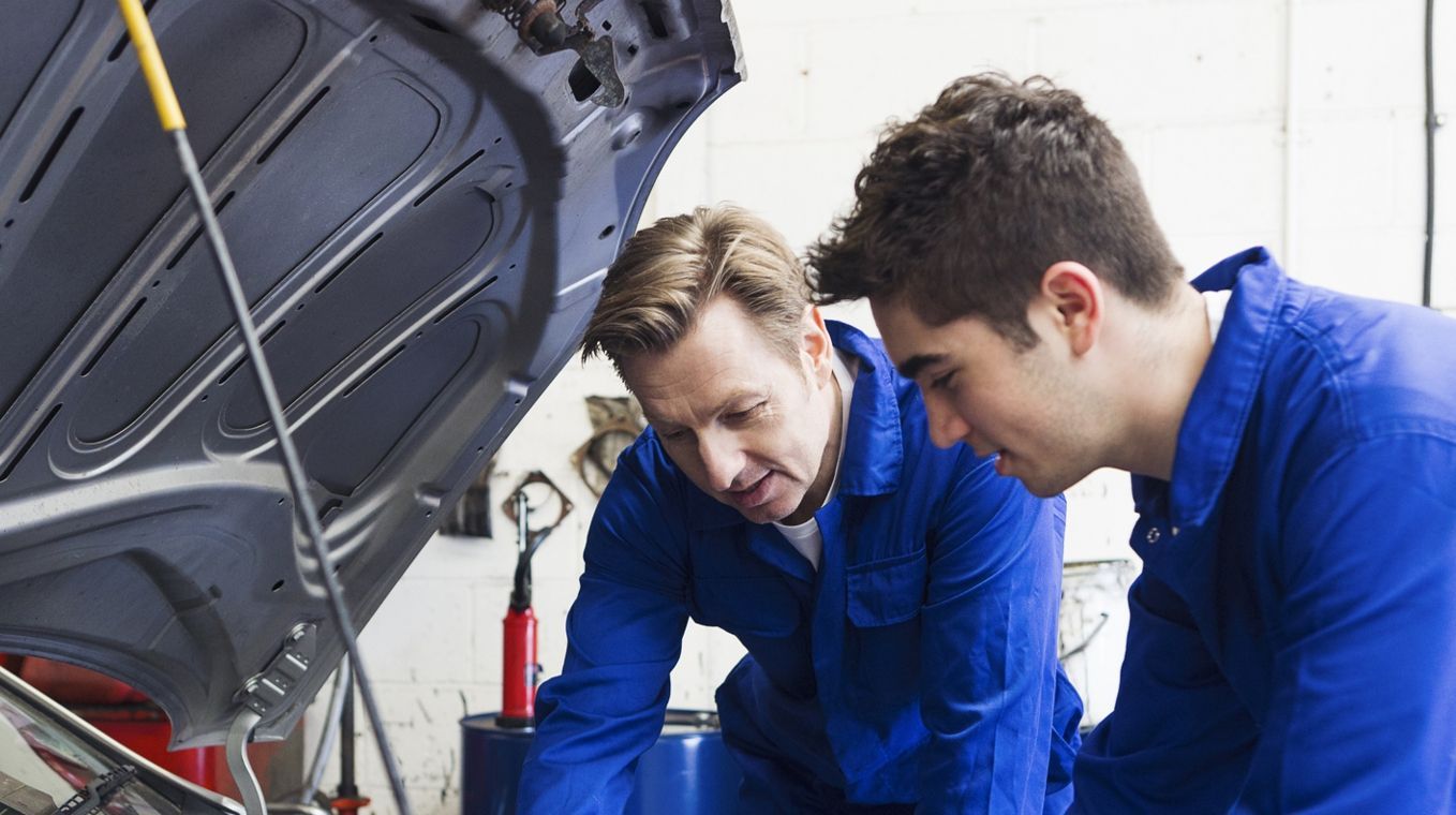 Zwei Männer in blauen Uniformen arbeiten in einer Werkstatt an einem Automotor.