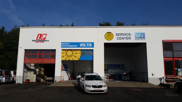 Ein weißes Gebäude mit verschiedenen Schildern, darunter 'LP Automobile', 'Ingenieur & Sachverstaendigenbuero', 'VOLTIS' und 'TUEV Hessen Service Center'. Vor dem Gebäude ist auch ein Auto geparkt.