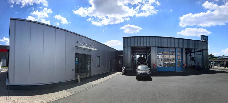 Ein modernes Gebäude mit einem blauen Schild, auf dem "TUEV Hessen" steht, neben einer Tankstelle, vor der ein Auto geparkt ist.