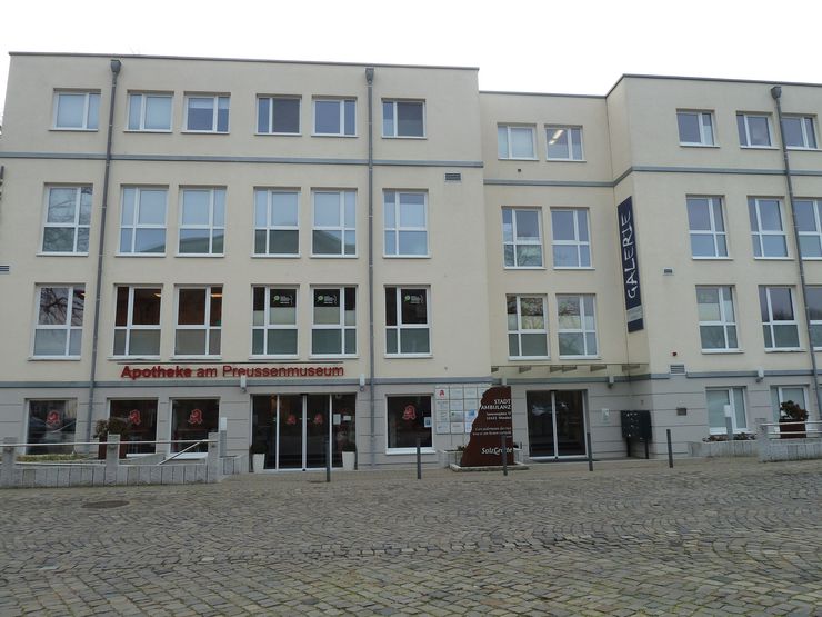 Ein mehrstöckiges Gebäude mit mehreren Fenstern, einem Schild mit der Aufschrift "Apotheke am Preussenmuseum" und einem Schild mit der Aufschrift "Galerie" auf der rechten Seite.