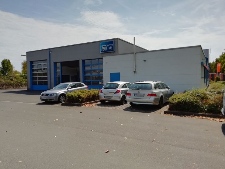 Ein Gebäude mit einem blauen Schild mit der Aufschrift "TUEV Hessen", eine Garage mit drei geparkten Autos und ein paar Bäume im Hintergrund.
