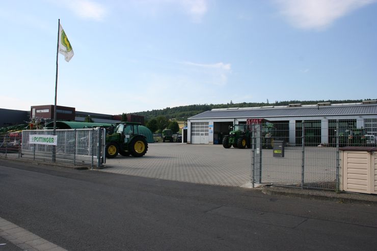 Ein Ausstellungsraum für Traktoren, vor dem ein grüner Traktor geparkt ist, ein weißes Gebäude mit grünem Dach und eine Flagge mit einem gelb-weißen Emblem.