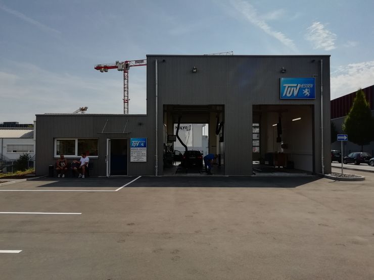 Eine moderne Autowerkstatt mit einem blauen Schild mit der Aufschrift 'TUEV HESSEN', ein paar Autos drinnen und ein paar Leute draußen.