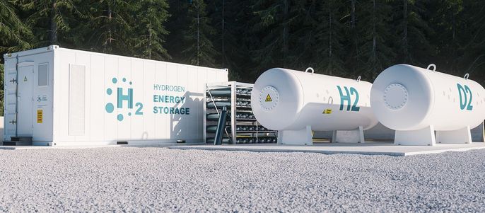 Eine moderne Anlage mit einem weißen Container mit der Aufschrift "Hydrogen Energy Storage" und zwei großen zylindrischen Tanks mit der Aufschrift "H2". Die Anlage ist von Bäumen umgeben, und es sind Sicherheitsschilder vorhanden.