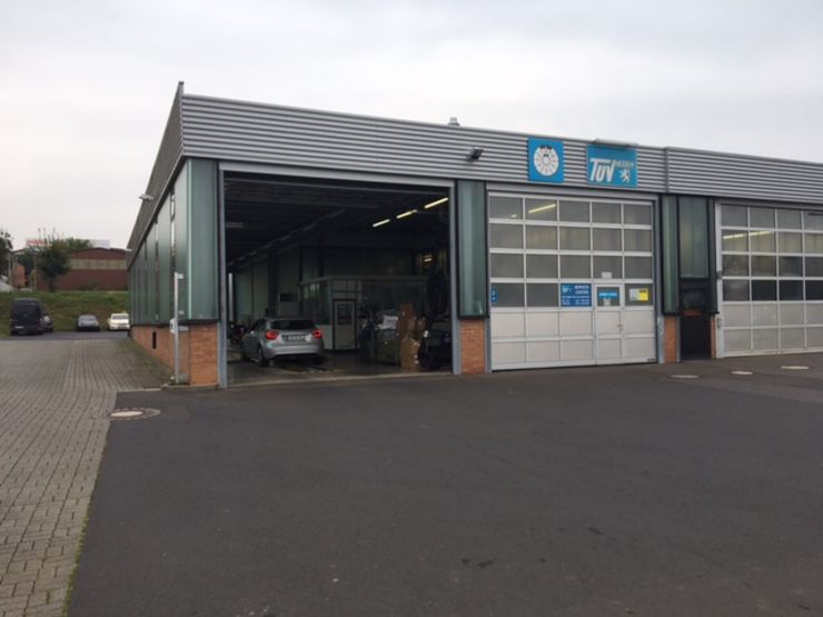Eine Garage mit einem blauen Schild mit der Aufschrift "TÜV Hessen", ein Auto darin und ein Parkplatz mit einigen Autos.