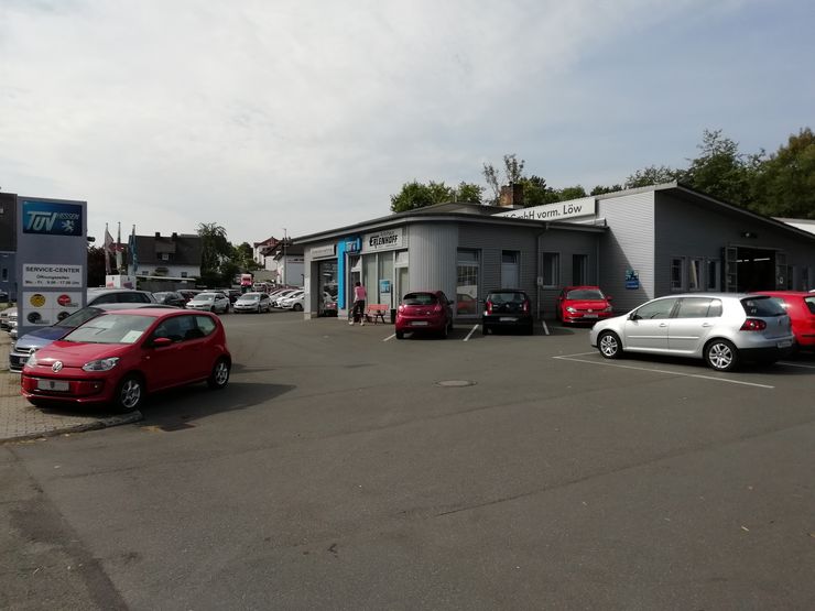Ein Autohaus mit verschiedenen Fahrzeugen, die draußen geparkt sind, darunter ein roter Volkswagen, ein silberner Volkswagen und ein blaues Auto. Außerdem gibt es ein Schild mit dem Logo von "TUEV Hessen" und ein weiteres Schild mit dem Namen des Autohauses.