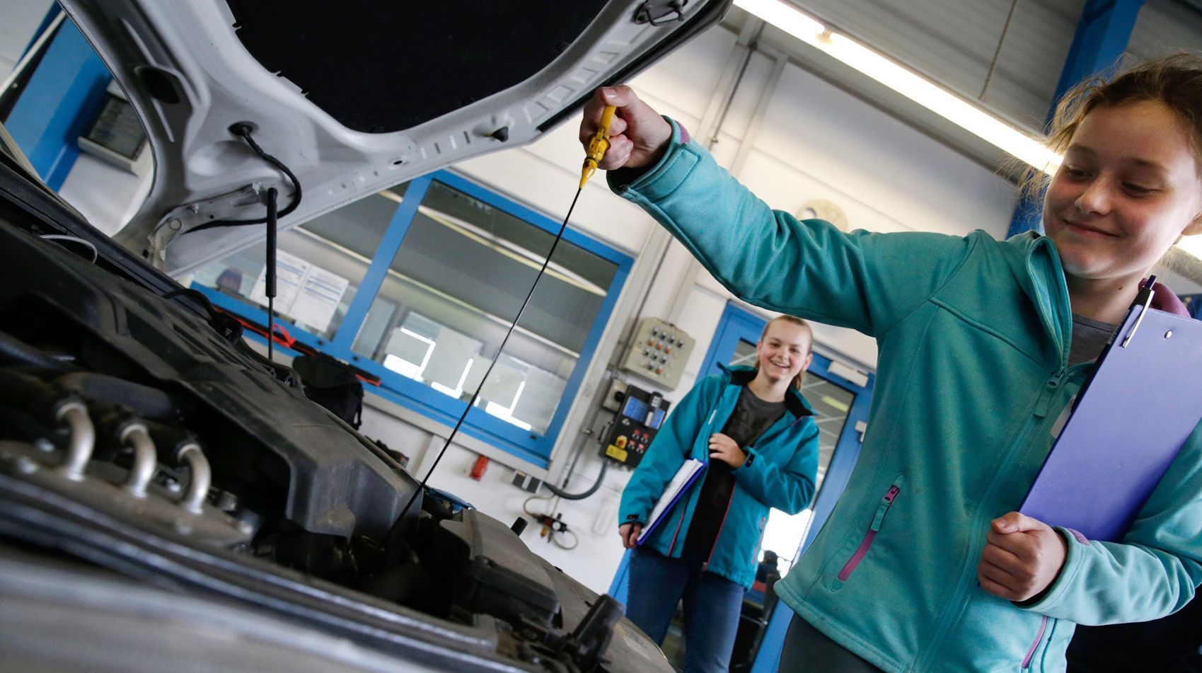 Ein junges Mädchen in einer blauen Jacke prüft den Ölstand eines Automotors in einer Werkstatt.
