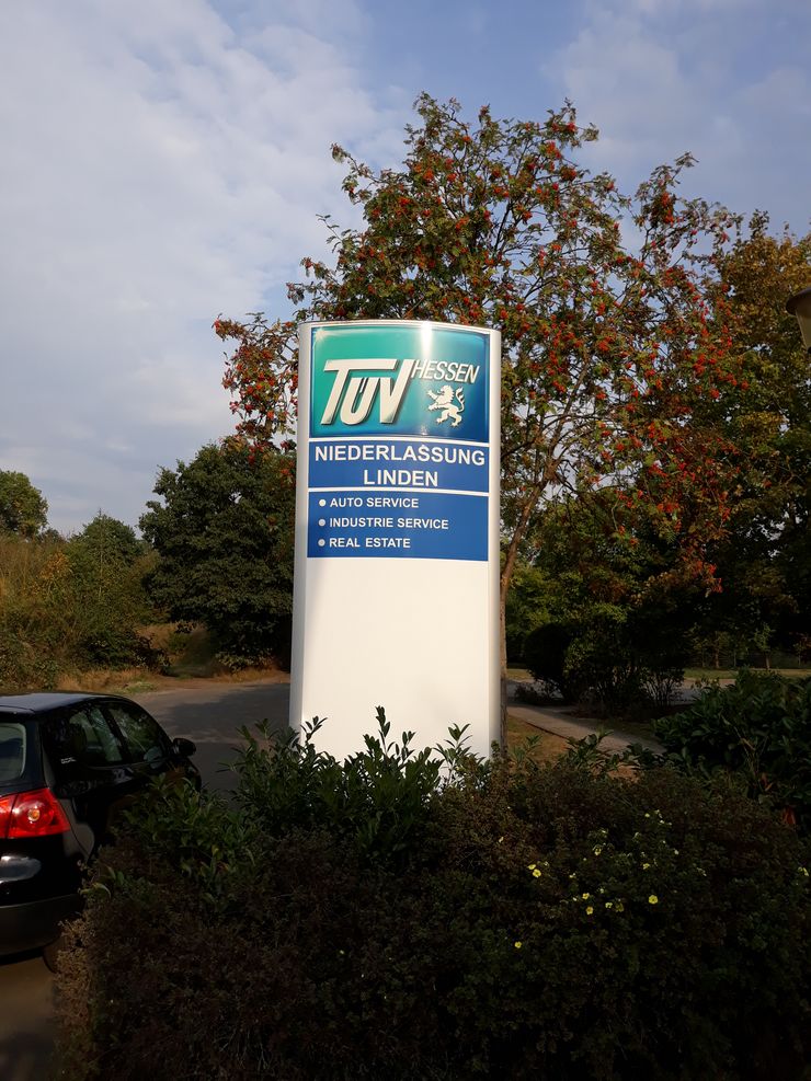Ein Schild für "TUEV Hessen" mit einem Richtungspfeil, der auf "Niederlassung Linden" zeigt. Das Schild bietet Informationen über den TUV-Service, einschließlich "Auto Service", "Industrie Service" und "Immobilien".