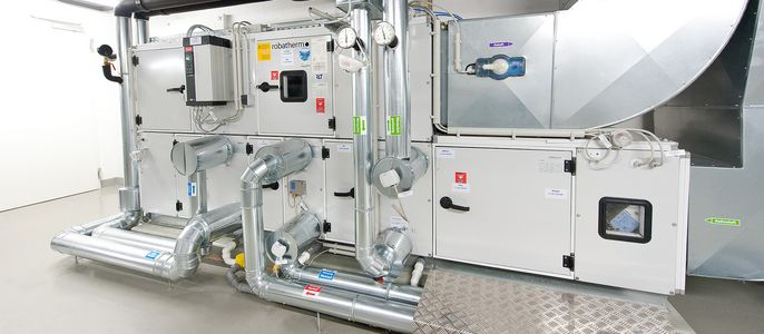 Ein komplexes mechanisches System, überwiegend in Silber und Weiß gehalten, mit verschiedenen Rohren, Ventilen und Schaltern, in einem Raum mit weißem Boden und weißen Wänden.