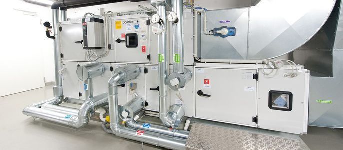 Ein komplexes mechanisches System, überwiegend in Silber und Weiß gehalten, mit verschiedenen Rohren, Ventilen und Schaltern, in einem Raum mit weißem Boden und weißen Wänden.