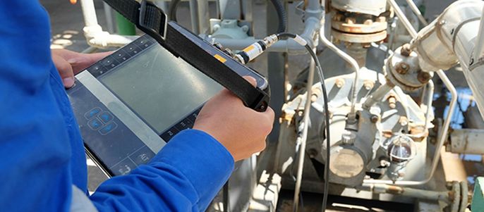 Eine Person in blauer Kleidung inspiziert mit einem Tablet-Gerät Maschinen, im Hintergrund sind verschiedene Rohre und Ventile zu sehen.