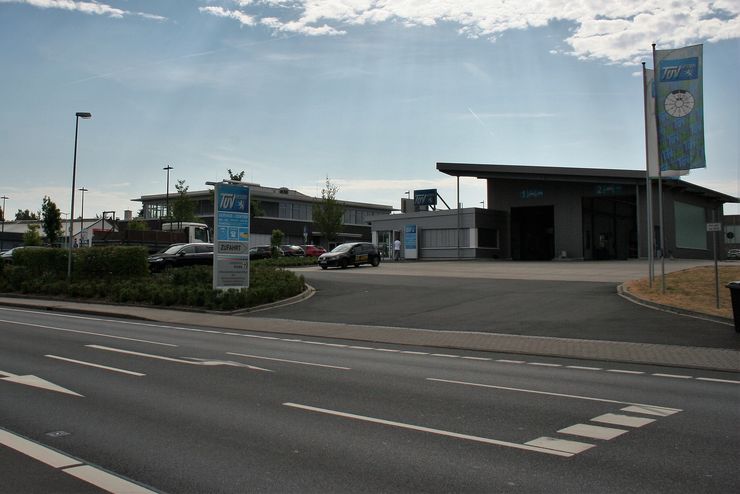 Eine moderne Autotankstelle mit einem Schild mit der Aufschrift "ZUFHART" und einem weiteren Schild mit der Aufschrift "TUEV Hessen". Draußen sind mehrere Autos geparkt, und der Himmel ist klar mit ein paar Wolken.