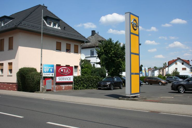 Eine Straßenszene mit einem Gebäude, einem Auto, einem Schild und einem hohen Mast mit dem Logo von TUEV Hessen.