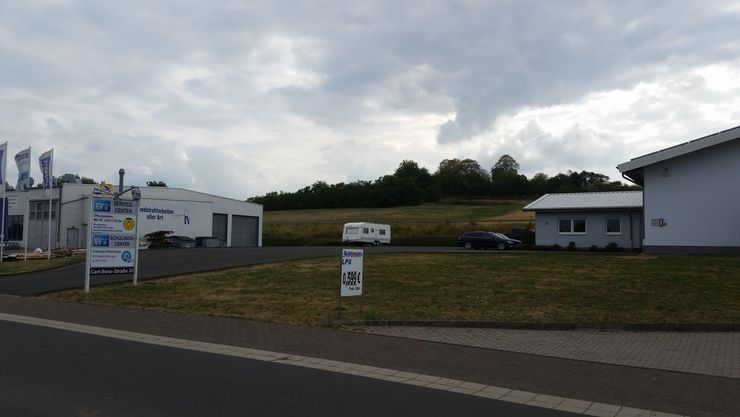 Ein Gewerbegebiet mit einem Gebäude mit der Aufschrift "TUEV Hessen SERVICE-CENTER" und einem weiteren mit der Aufschrift "Industrietechnik". Es gibt mehrere Fahnen, ein Auto, einen Anhänger.