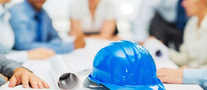 Eine Gruppe von Menschen in einer Besprechung, mit einem blauen Schutzhelm und aufgerollten Bauplänen auf dem Tisch.