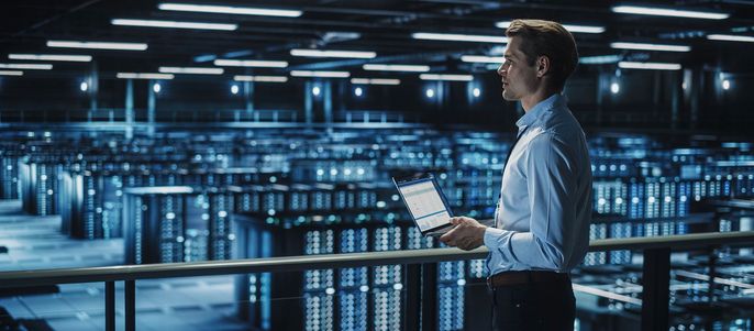 Ein Mann in einem blauen Hemd steht in einem Rechenzentrum mit zahlreichen Servern und hält einen Laptop, auf dem Daten angezeigt werden.