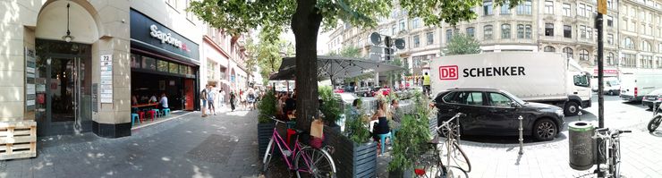 Eine belebte städtische Straße mit verschiedenen Einrichtungen, darunter ein Café, ein Geschäft und ein Lastwagen mit dem Logo "DB SCHENKER". Außerdem gibt es Spaziergänger, abgestellte Fahrräder und schattenspendende Bäume.