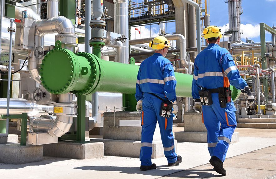 Two engineers in blue overalls and yellow hard hats walk along a bank of pipes at a refinery