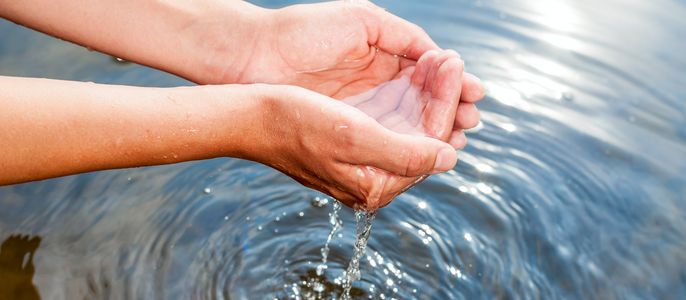 Die Hände einer Person, die klares Wasser halten, das im Wasser darunter Wellen verursacht.