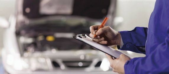 Eine Person in blauer Uniform, die ein Klemmbrett und einen Stift in der Hand hält, inspiziert den Motor eines Autos.