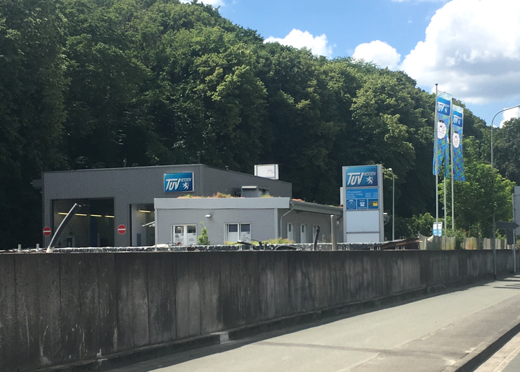 Ein modernes Gebäude mit dem "TUEV Hessen"-Logo, umgeben von Grün, mit einer Straße und ein paar Fahnen im Hintergrund.