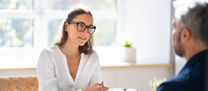 Eine Frau mit Brille und weißer Bluse unterhält sich mit einem Mann, beide sitzen in einem gut beleuchteten Raum mit einem Fenster.