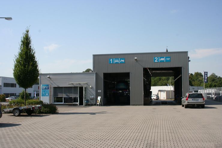 Ein modernes Gebäude mit zwei Garagentoren, einem Schild mit der Aufschrift "TUEV Hessen" und einigen davor geparkten Fahrzeugen.