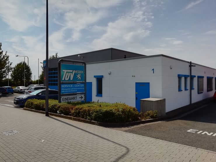 Ein Gebäude mit einem Schild mit der Aufschrift "TUEV HESSEN" und "SERVICE-CENTER" sowie einer Telefonnummer. Das Gebäude ist weiß und blau gestrichen, und vor dem Gebäude sind Autos geparkt.