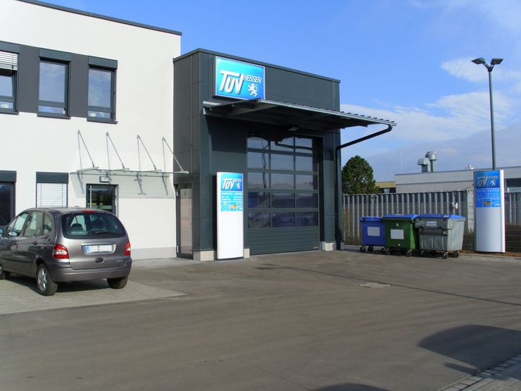 Ein Gebäude mit einem "TUEV Hessen"-Schild, ein vor dem Gebäude geparktes Auto und verschiedene Mülltonnen.