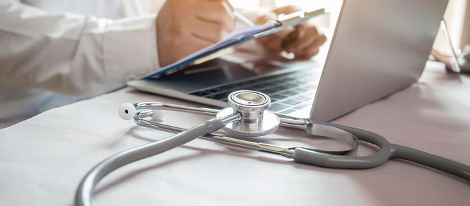 Eine medizinische Fachkraft, vermutlich ein Arzt, benutzt einen Laptop und hält ein Klemmbrett. Auf dem Tisch neben dem Laptop befindet sich ein Stethoskop.
