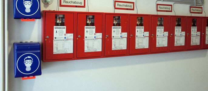 Eine Reihe roter Schränke mit der Aufschrift "Rauchabzug", in denen sich verschiedene Geräte befinden, begleitet von blauen Schränken mit dem Symbol einer Person, die Kopfhörer trägt.