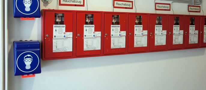 Eine Reihe roter Schränke mit der Aufschrift "Rauchabzug", in denen sich verschiedene Geräte befinden, begleitet von blauen Schränken mit dem Symbol einer Person, die Kopfhörer trägt.