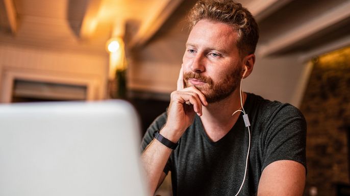Ein Mann mit lockigem Haar, der ein schwarzes Hemd trägt und mit Kopfhörern in den Ohren konzentriert auf einen Laptop-Bildschirm schaut.