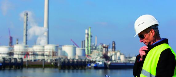 Ein Mann in Sicherheitsweste und Schutzhelm, der mit einem Walkie-Talkie spricht, vor einem industriellen Hintergrund mit großen Tanks und Schornsteinen.