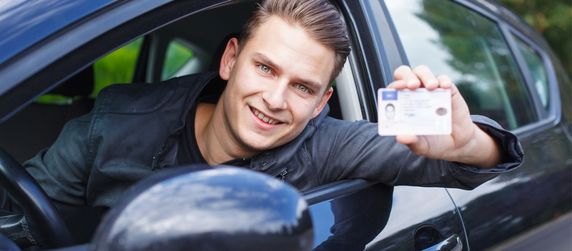 Ein junger Mann zeigt, aus einem Auto lehnend, seinen Führerschein.