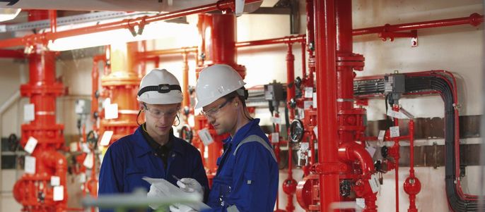 Zwei Arbeiter in einer Fabrikumgebung, umgeben von roten Rohren und Maschinen, diskutieren über ein Dokument.