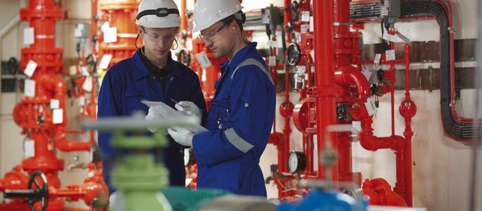 Zwei Arbeiter in einer Fabrikumgebung, umgeben von roten Rohren und Maschinen, diskutieren über ein Dokument.