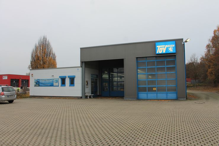 Ein modernes Gebäude mit einem blauen "TUEV Hessen"-Logo an der Fassade, das auf einem offenen Parkplatz mit einigen Autos und Bäumen im Hintergrund steht.