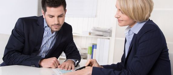 Eine Frau und ein Mann sprechen an einem Schreibtisch miteinander. Ein Mobiltelefon und Papieren liegen auf dem Tisch.