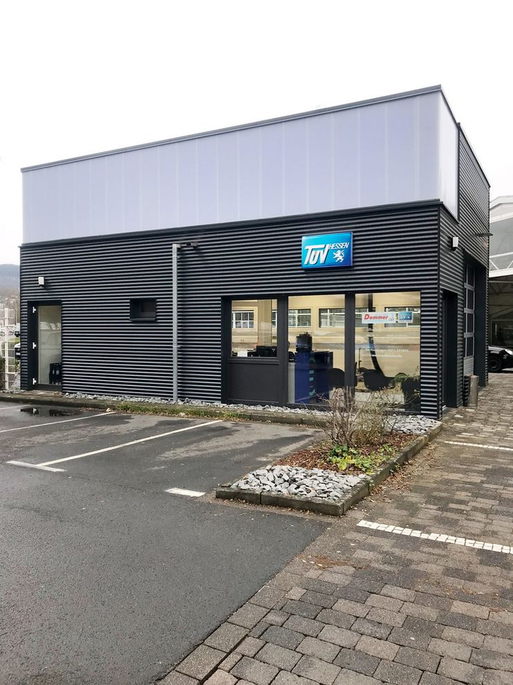Ein modernes Gebäude mit dunkelgrauer Fassade, einem blauen Schild mit der Aufschrift 'TUEV Hessen' und ein paar geparkten Autos.