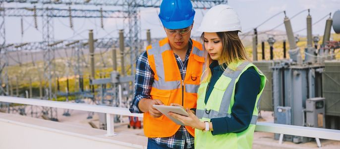 Zwei Arbeiter, ein Mann und eine Frau, die Sicherheitswesten und Helme tragen, untersuchen ein Tablet-Gerät in einer Außenumgebung mit elektrischen Geräten im Hintergrund.