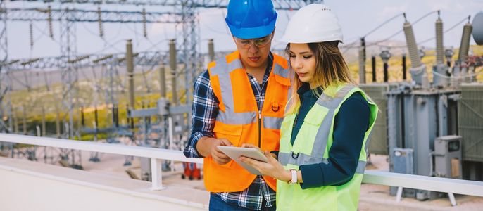 Zwei Arbeiter, ein Mann und eine Frau, die Sicherheitswesten und Helme tragen, untersuchen ein Tablet-Gerät in einer Außenumgebung mit elektrischen Geräten im Hintergrund.