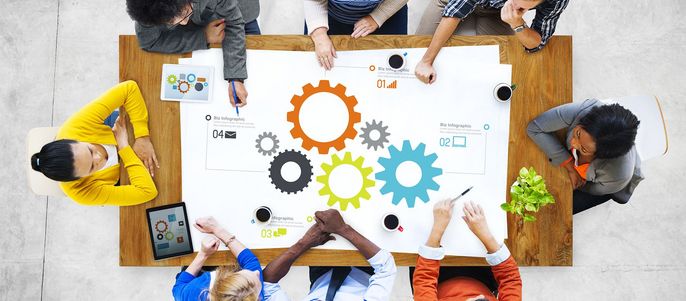 Eine Gruppe von Menschen, die an einem Tisch sitzen und an einem Projekt arbeiten. Sie sind von großen bunten Zahnrädern umgeben, einige halten Stifte und Tablets, und auf dem Tisch sind verschiedene Symbole und Zahlen zu sehen.