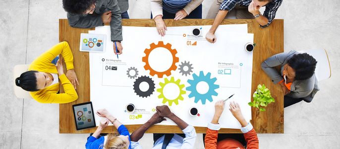 Eine Gruppe von Menschen, die an einem Tisch sitzen und an einem Projekt arbeiten. Sie sind von großen bunten Zahnrädern umgeben, einige halten Stifte und Tablets, und auf dem Tisch sind verschiedene Symbole und Zahlen zu sehen.