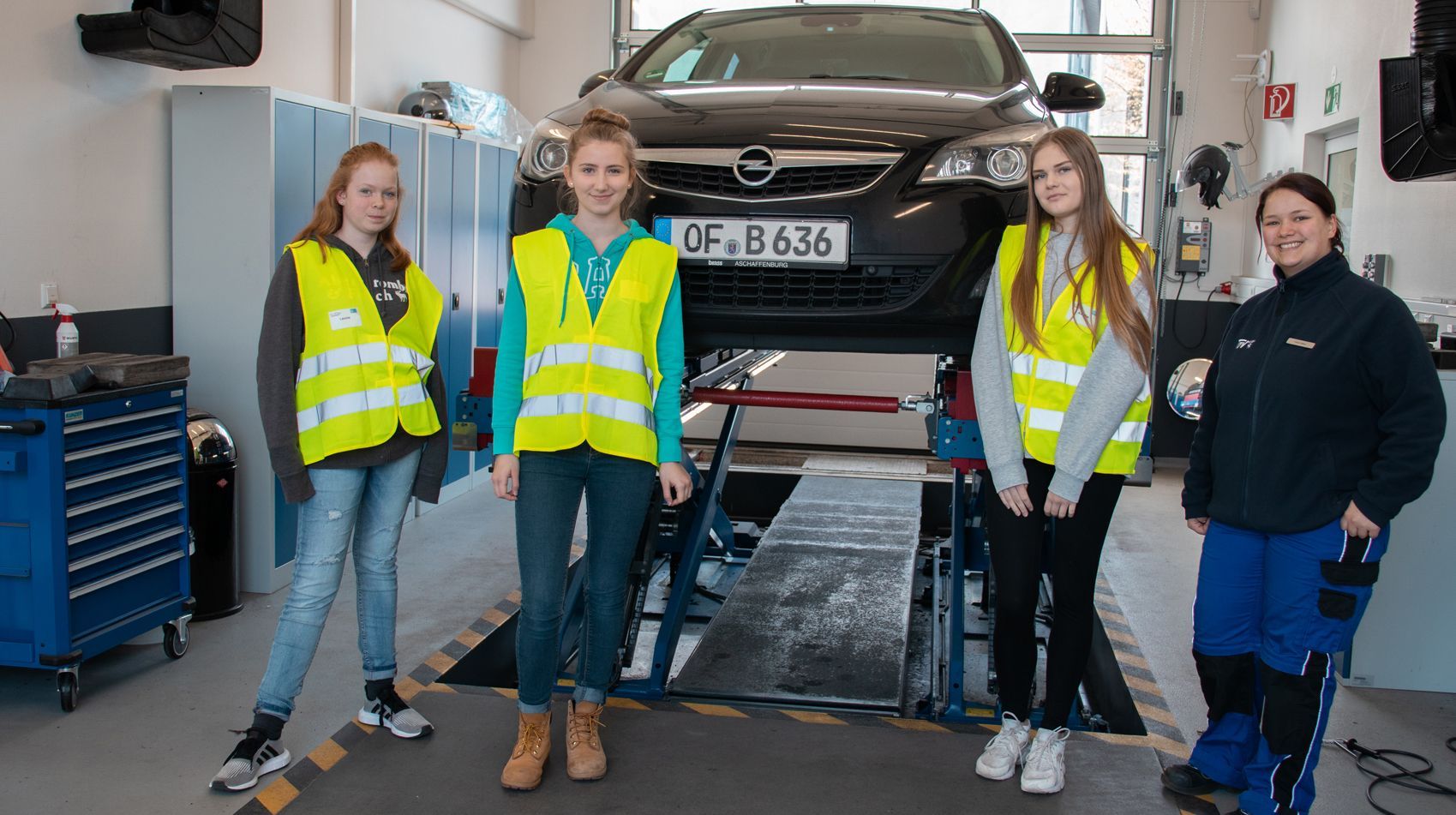 Vier Personen in einer Werkstatt, mit einem Auto auf einer Hebebühne im Hintergrund. Sie tragen reflektierende Westen und stehen vor verschiedenen Werkzeugen und Geräten.