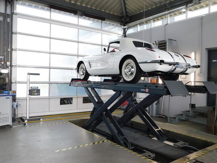 Ein weißer Oldtimer auf einer Hebebühne in einer Werkstatt, um den herum verschiedene Werkzeuge und Geräte liegen.