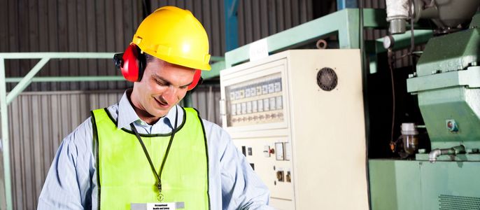 Ein Mann, der einen gelben Schutzhelm und eine grüne Weste trägt, steht in einer Fabrikhalle und schreibt etwas auf ein Klemmbrett.